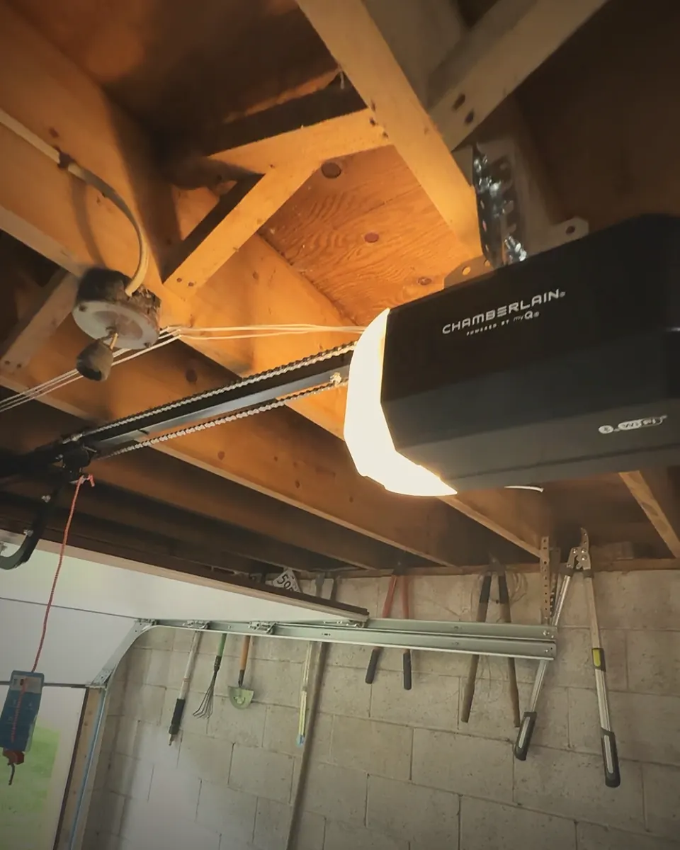 Black Chamberlain myQ Wi-Fi chain-drive garage door opener mounted to the ceiling joists of a customer's garage, with its light on and the rail running to the door below